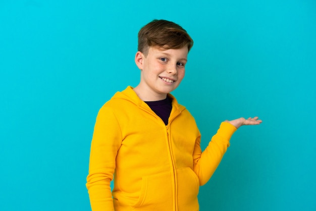 Little redhead boy isolated on blue wall holding copyspace imaginary on the palm to insert an ad