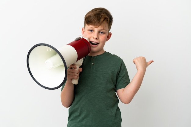 Little redhead boy isolated on white background shouting through a megaphone and pointing side