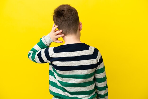 Little redhead boy isolated on yellow background in back position and thinking