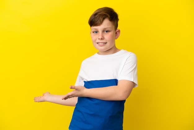 Little redhead boy isolated on yellow background extending hands to the side for inviting to come