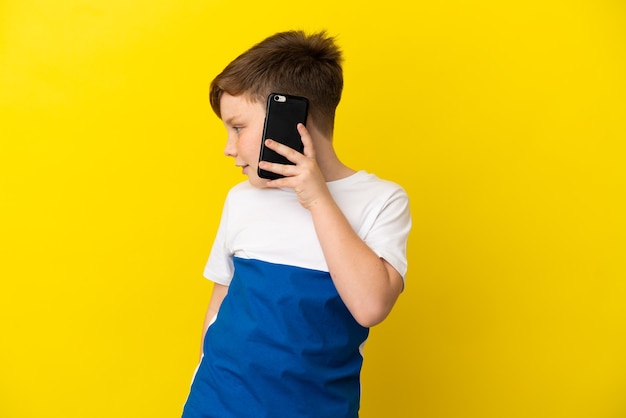 Little redhead boy isolated on yellow background keeping a conversation with the mobile phone with someone