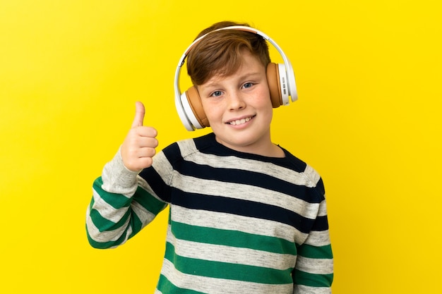 Little redhead boy isolated on yellow background listening music and with thumb up