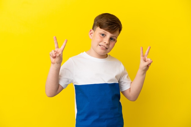 Little redhead boy isolated on yellow background showing victory sign with both hands