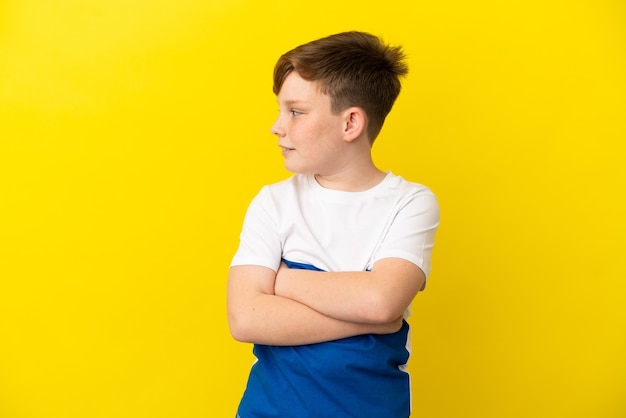 Little redhead boy isolated on yellow background with arms crossed and happy