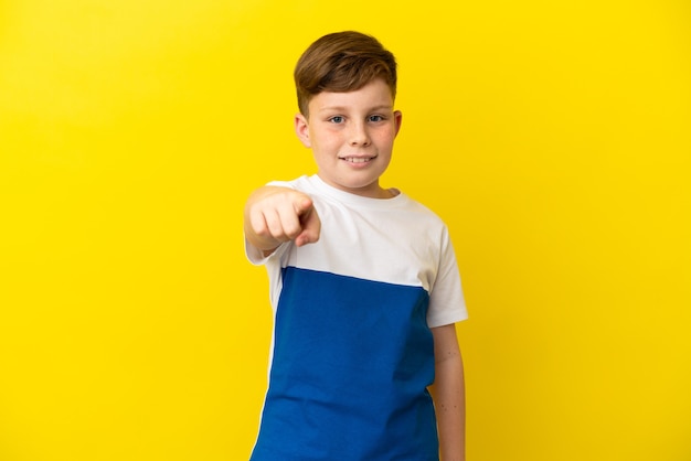 Little redhead boy isolated on yellow surface surprised and pointing front
