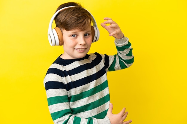 Little redhead boy isolated on yellow wall listening music and doing guitar gesture