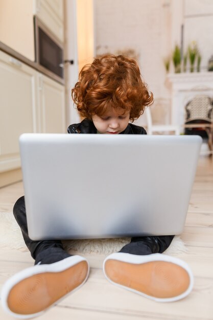 Little redhead boy uses a laptop while sitting on the floor at home