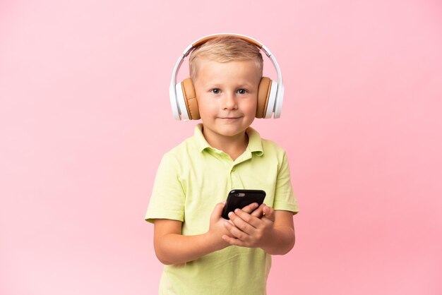 Little Russian boy isolated on pink background listening music and pointing to the front