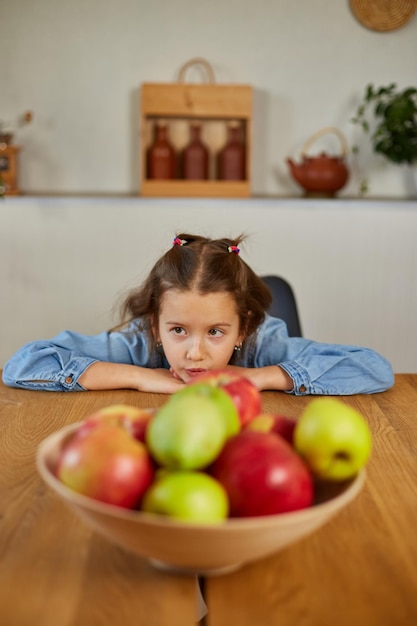 Little sad girl look on bowl with fruits in the kitchen at home