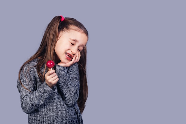 Piccola ragazza triste con un lecca-lecca rosso tra le mani, i cui denti fanno male. il concetto di sviluppo della carie dovuto all'abuso di caramelle. isolato su superficie grigia