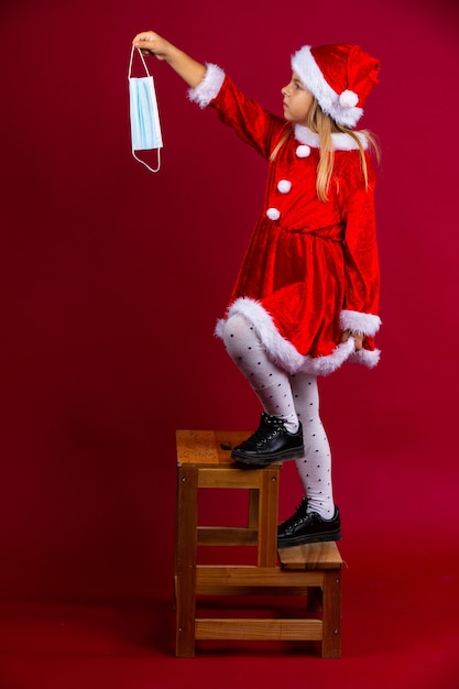 The little santa girl climbs a ladder and wants to decorate something with a mask, isolated on red