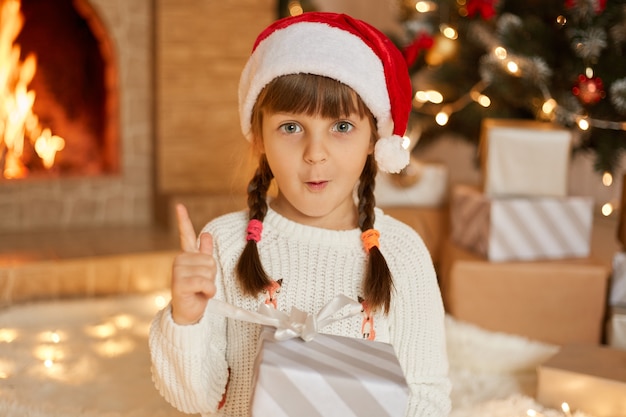 Little Santa girl in red hat and white jumper, posing indoor in festive room