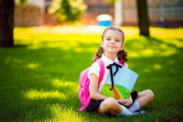 レッスンの後草の上に座っているピンクのバックパックと小さな学校の女の子と本を読んだり、レッスン、思考のアイデア、教育、学習の概念を勉強します。