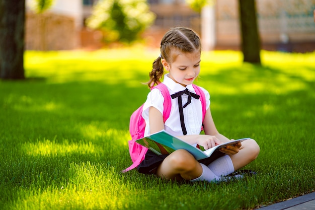 Little school girl with pink backpack sitting on grass after lessons and read book or study lessons,