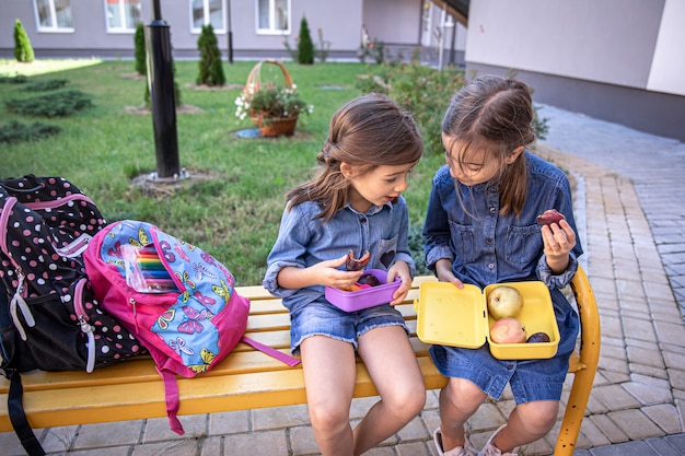 Little school girls sitting on bench in school yard and eating from lunch boxes.