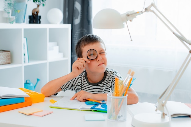 Little schoolboy boy is sitting at home at the table and solving homework looking through a magnifying glass . Back to school, preparation, home schooling.