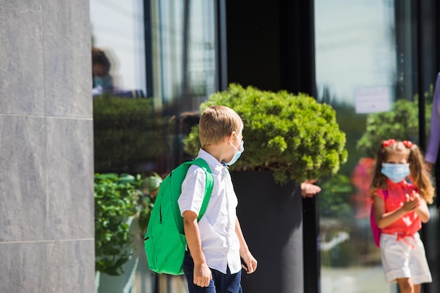 Il piccolo scolaro che indossa una maschera per il viso in piedi fuori vicino alla scuola il ragazzo che aspetta i suoi compagni di scuola