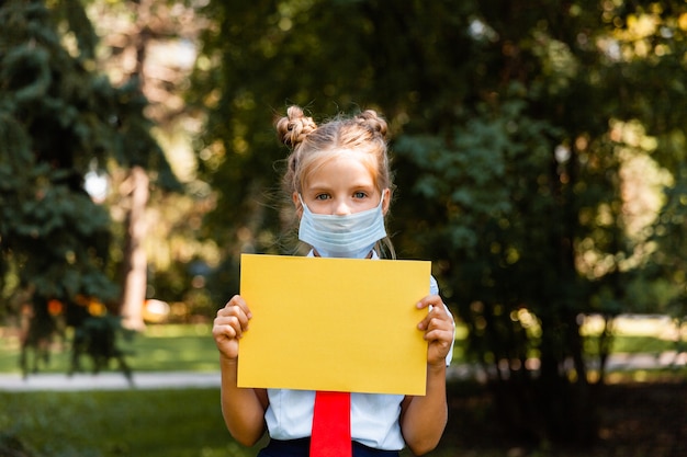 Little schoolgirl in a medical mask sits in the park on the grass and has fun.