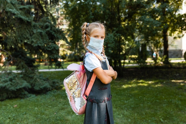 Little schoolgirl in a medical mask sits in the park on the grass and has fun.