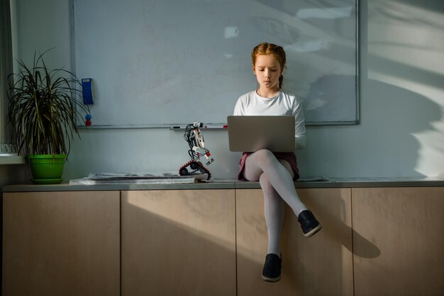 little schoolgirl using laptop on table at classroom with diy robot