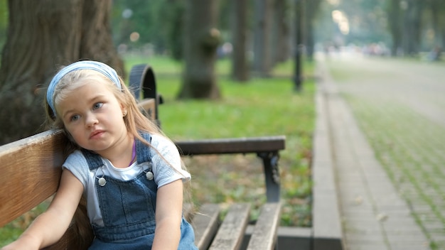 Little serious child girl sitting alone on a bench in summer park.