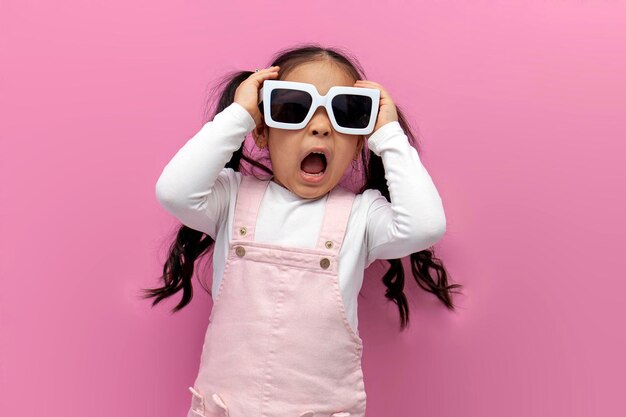 little shocked preschool girl in white sunglasses and pink sundress screams and is surprised