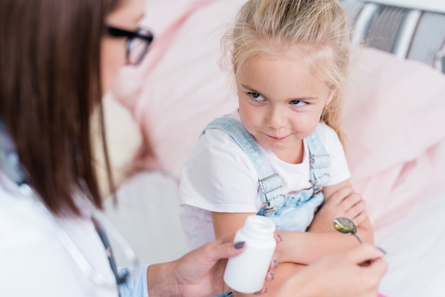 Little sick capricious girl with crossed arms and reluctant expression looking at doctor giving her pills in hospital