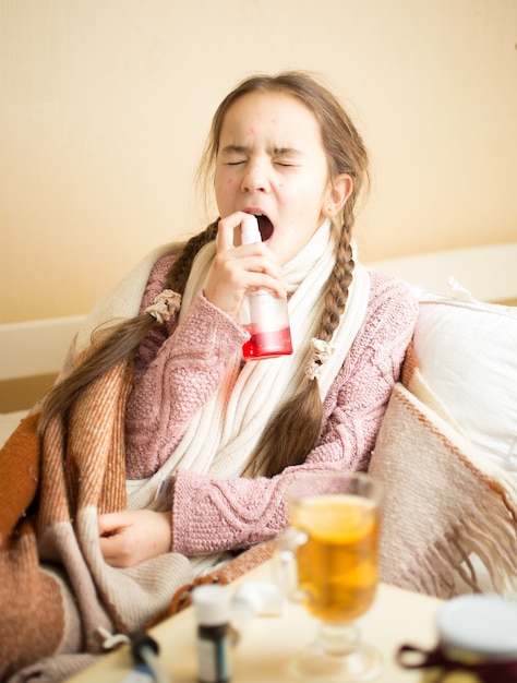 Little sick girl lying in bed and using throat spray