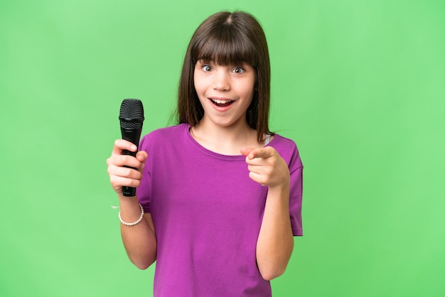 Маленькая певица, поднимающая микрофон на изолированном фоне, удивлена и указывая вперед