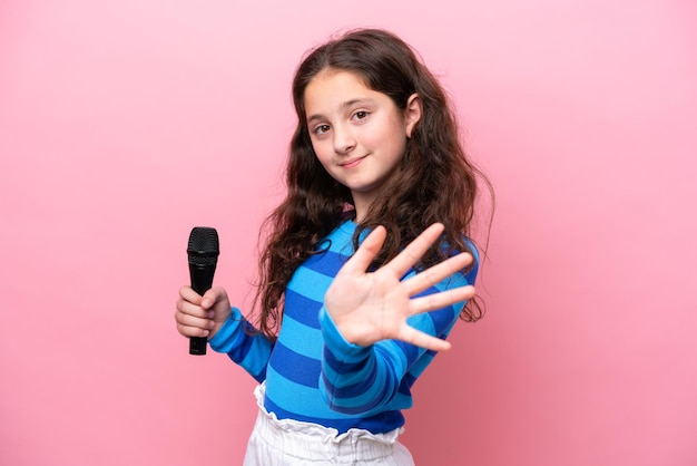 Little singer girl picking up a microphone isolated on pink background saluting with hand with happy expression