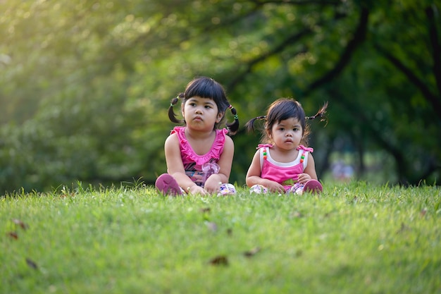 Little sister play and sit on the green lawn in park. Cute girls relax in park.