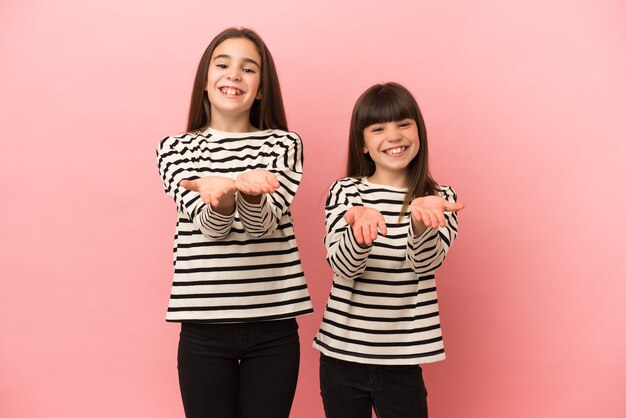 Little sisters girls isolated on pink background holding copyspace imaginary on the palm to insert an ad