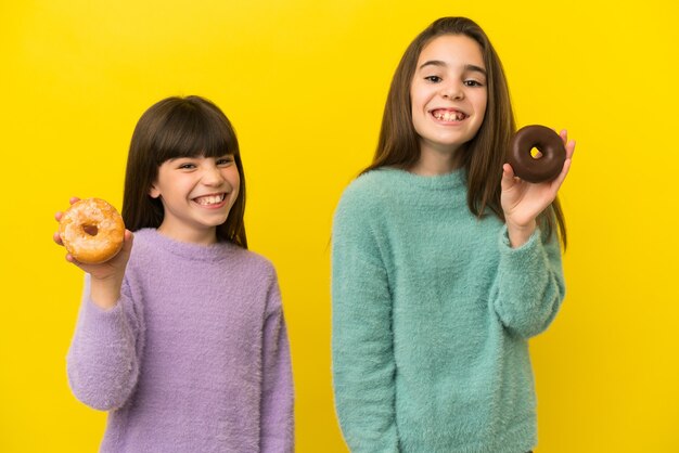 Little sisters isolated on yellow background holding donuts with happy expression