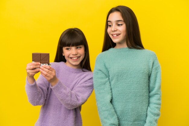 Little sisters isolated on yellow background taking a chocolate tablet and happy