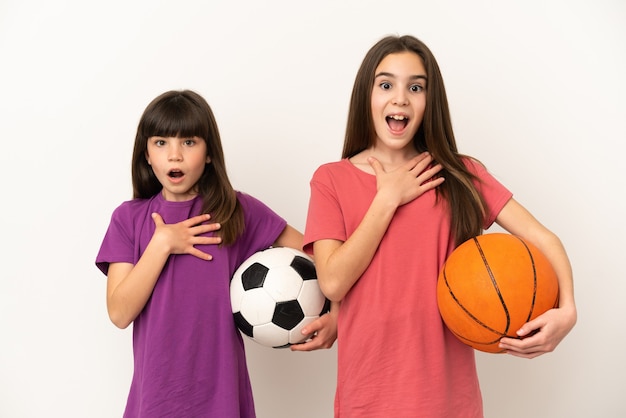 흰색 배경에 격리된 축구와 농구를 하는 어린 자매들은 오른쪽을 보고 놀라고 충격을 받았습니다.