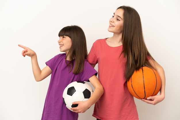 Little sisters playing football and basketball isolated on white background presenting an idea while looking smiling towards