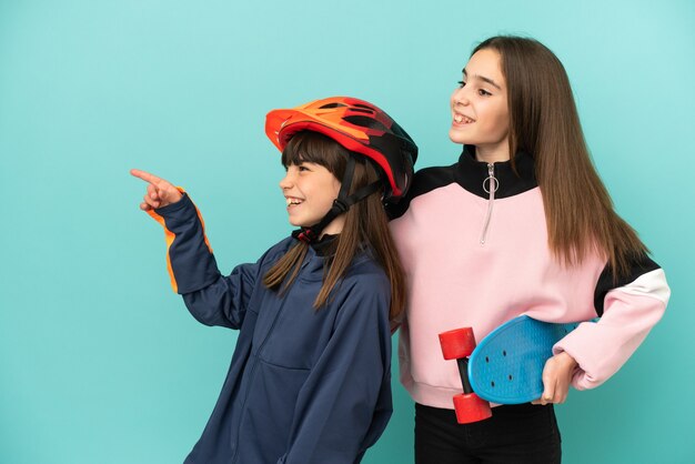 Little sisters practicing cycling and skater isolated on blue background presenting an idea while looking smiling towards