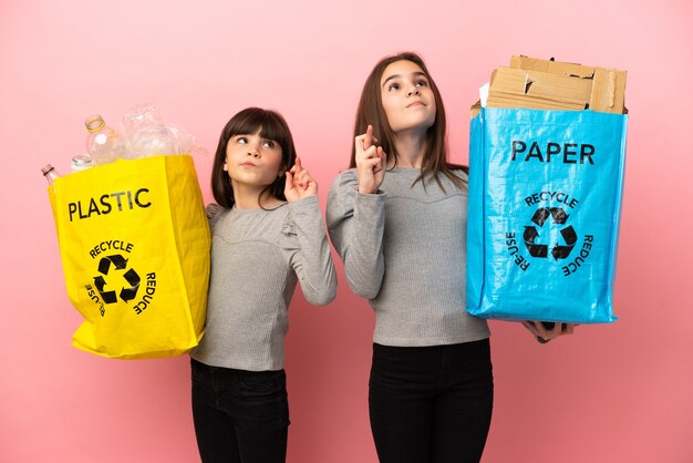 Little sisters recycling paper and plastic isolated on pink background with fingers crossing and wishing the best