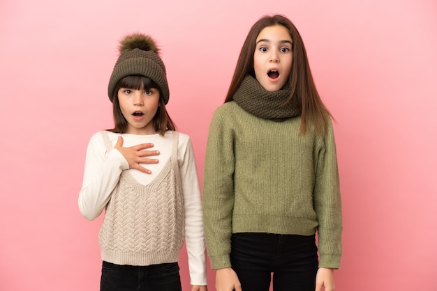 Little sisters wearing a winter clothes isolated on pink background with surprise and shocked facial expression