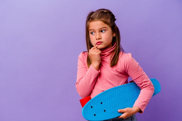 Little skater caucasian girl isolated on blue background relaxed thinking about something looking at a copy space.