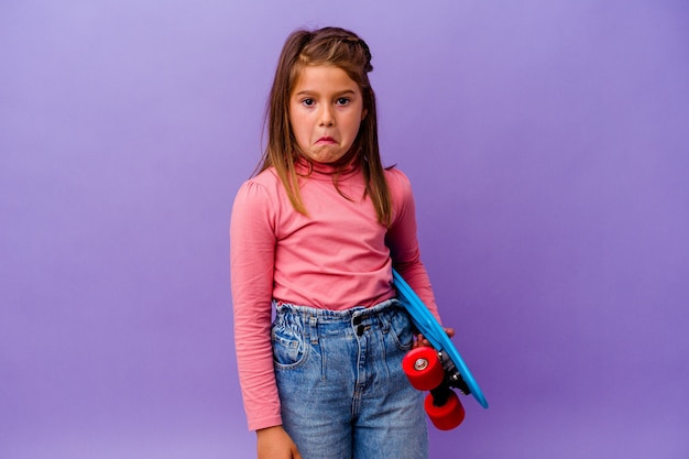 Little skater caucasian girl isolated on blue wall shrugs shoulders and open eyes confused