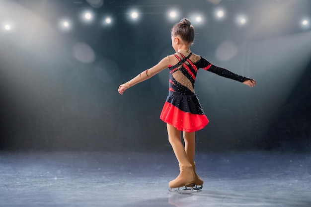 작은 스케이팅 선수는 아이스 경기장에서 빨간색과 검은색 드레스를 입고 링을 탄다