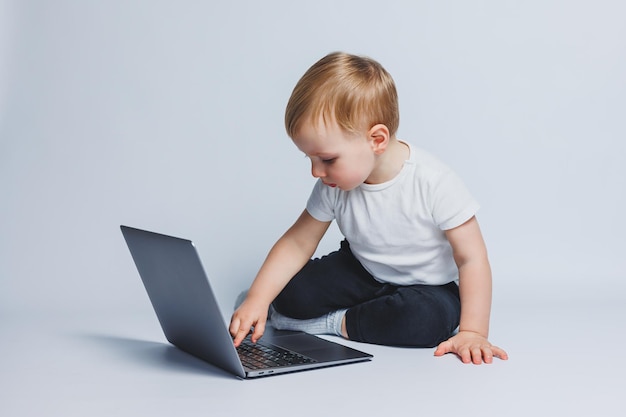 34세의 똑똑한 소년 흰색 배경에 노트북과 함께 앉아 흰색 티셔츠와 검은색 바지를 입은 아이가 노트북에 앉아 화면을 바라보고 있습니다. 현대 진보적인 아이들