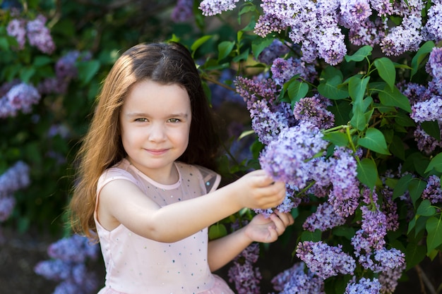Una bambina di sorriso è tra i cespugli di lillà nel giardino del tramonto, parco fiorito di primavera.