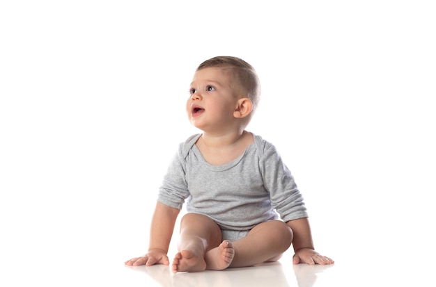 Little smiling baby in bodysuit sitting barefoot on the floor isolated on white