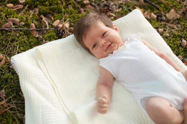 Little smiling baby in white clothing lying on the white blanket on the grass