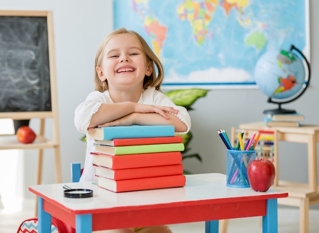 Маленькая улыбающаяся белокурая девушка сидит за белым столом и держит руки на книгах в просторном школьном классе
