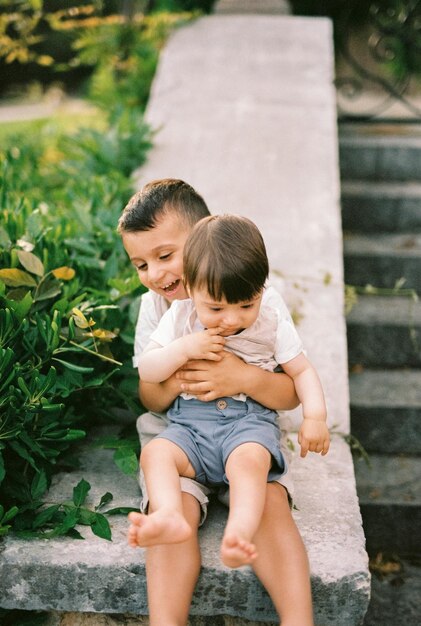 Foto piccolo ragazzo sorridente che tiene il fratello minore tra le braccia mentre è seduto su una ringhiera di pietra nel