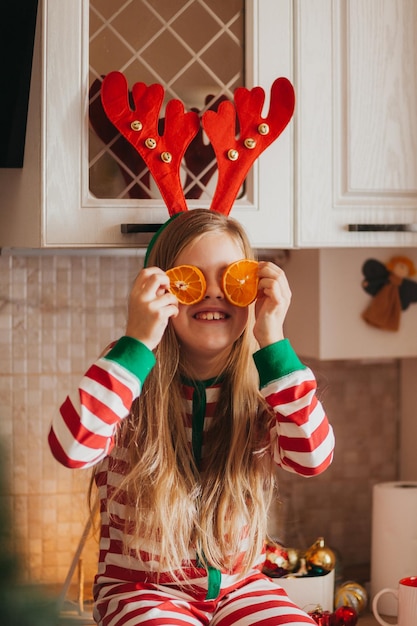 Маленькая улыбающаяся милая блондинка в пижаме и рогах держит половинки фруктов возле глаз. ребенок на кухне возле елки. рождество