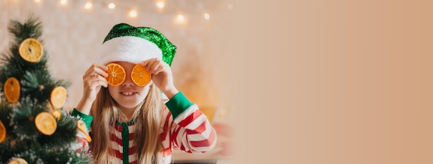 Little smiling cute blonde girl in pajamas and antlers holds fruit halves near her eyes child in the kitchen near the Christmas tree Christmas